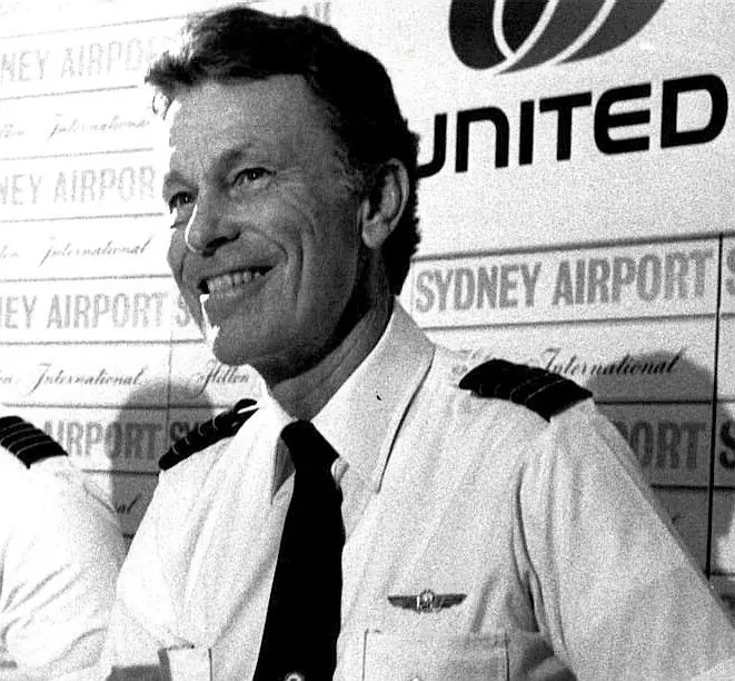 Black-and-white press photograph of a United Airlines captain in a white uniform shirt and dark tie, smiling, in front of a United and Sydney Airport backdrop.