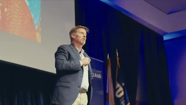 Stuart McClure speaking at Secure Miami, one hand on his chest, beside the event's banner and flags.