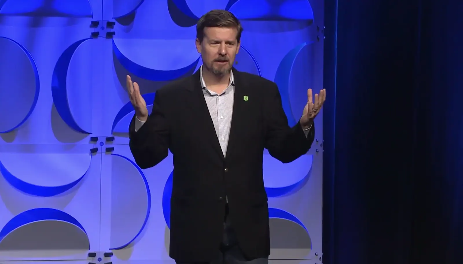 Stuart McClure on stage in a dark blazer, both palms open toward the audience, in front of a blue-lit wall of circles.